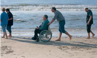 a picture of some people walking along the sand with a person in a wheelchair